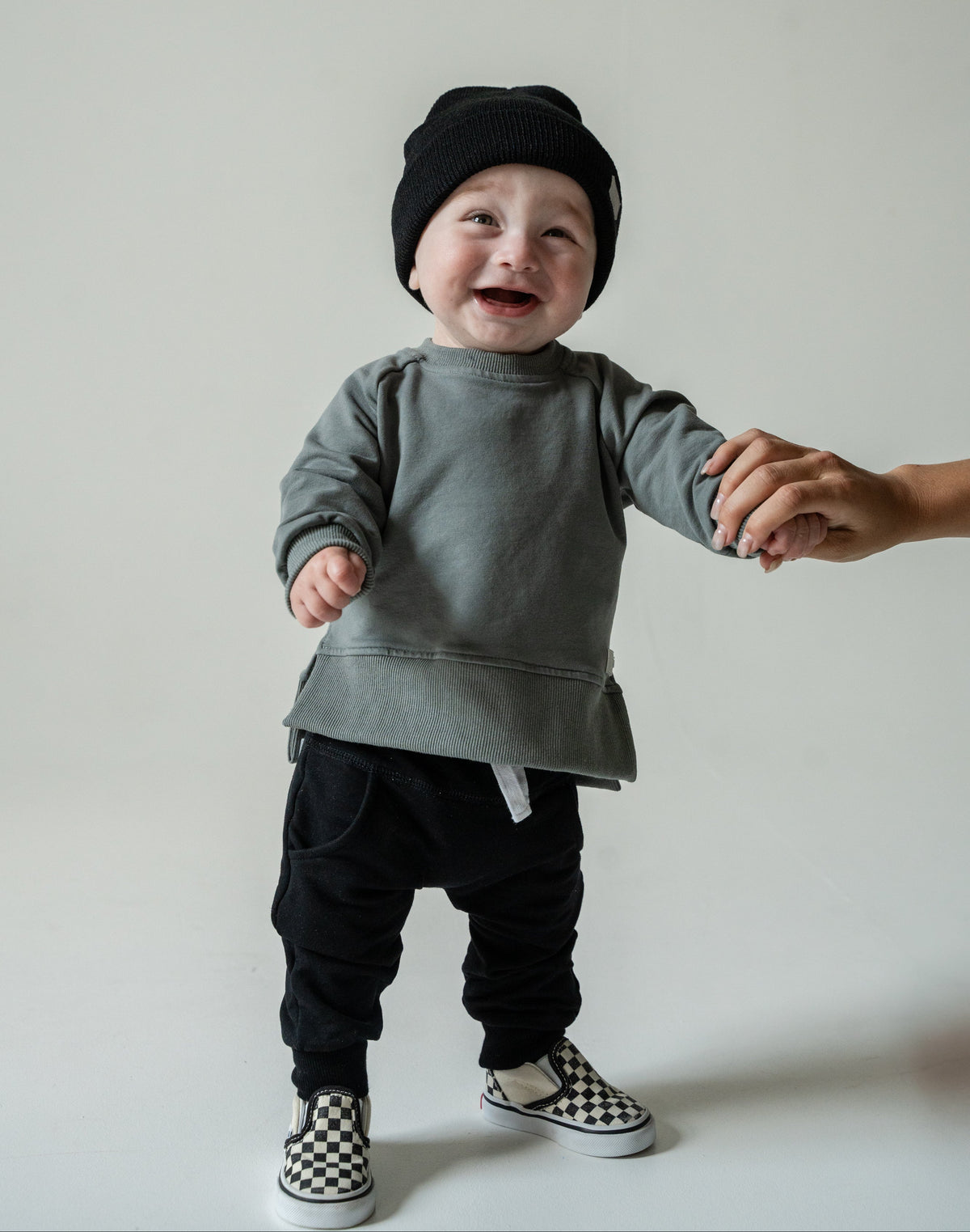 A baby wearing a gray long-sleeve top, black pants, a black beanie, and checkerboard slip-on shoes is smiling and holding an adult's hand against a plain light gray studio background.